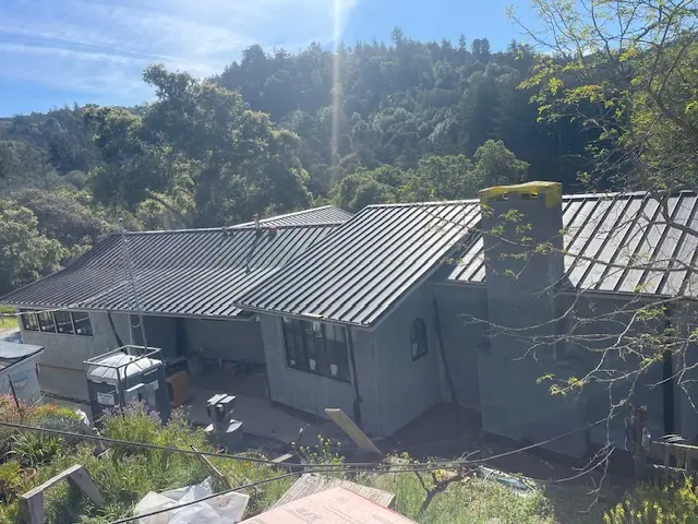 Dark standing-seam metal roof around a chimney on a hillside home overlooking wooded hills